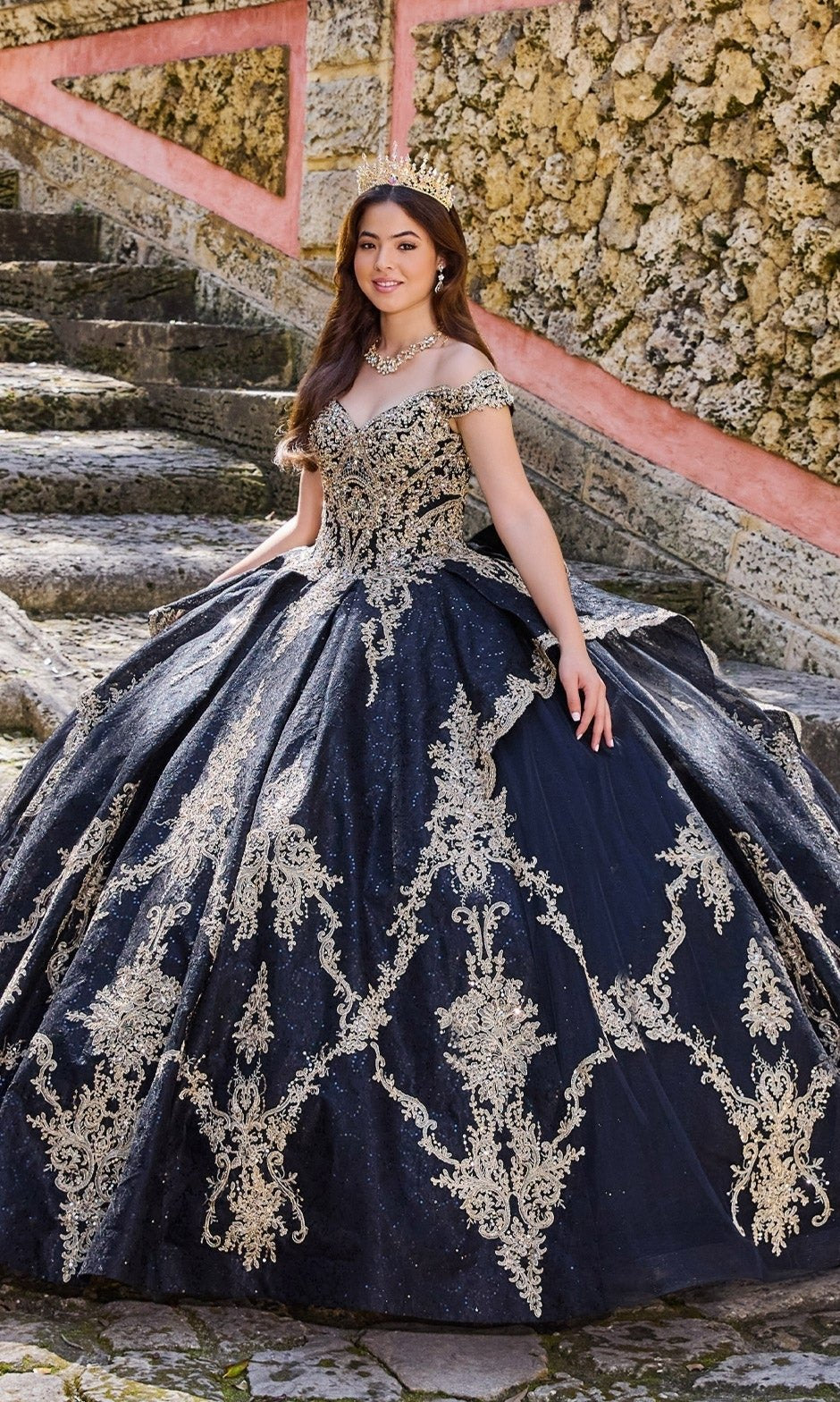 A young woman in the Quinceanera Dress PR30138 by Princesa, a navy blue and gold off-the-shoulder gown with glitter tulle and tiara, stands on stone steps outdoors.